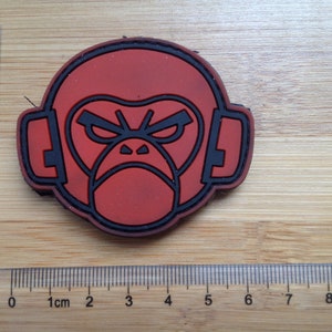 May include: A circular red and black rubber patch featuring a stylized monkey face wearing headphones. The patch is positioned next to a clear plastic ruler, showing a width of approximately 3.5 inches and a height of approximately 2.5 inches.