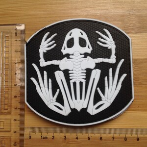 Pode incluir: Um patch preto e branco com um design de esqueleto de rã. O patch tem um fundo preto com um padrão texturizado e um esqueleto de rã branco no centro. O patch tem aproximadamente 7,6 cm de largura.