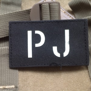 Puede incluir: Parche rectangular negro con las letras blancas "PJ". El parche está cosido sobre un fondo beige y verde oliva. Fabricado con tejido resistente, probablemente para fijar a bolsas o equipos.