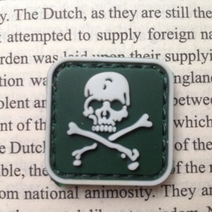 May include: A green fabric patch with a white skull and crossbones design. The patch is square and has a stitched border.