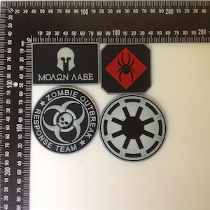 Puede incluir: Cuatro parches bordados en blanco y negro con diferentes diseños. Los parches están sobre una superficie blanca. Los parches incluyen una calavera y huesos cruzados con el texto "Zombie Outbreak Response Team", una araña negra sobre un diamante rojo, un símbolo blanco y negro con un círculo y ocho radios, y un casco blanco y negro con el texto "Molon Labe".
