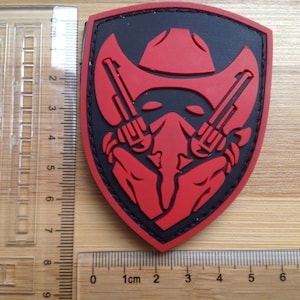 May include: Red and black embroidered patch featuring a skull wearing a cowboy hat and holding two pistols. The patch is shaped like a shield.