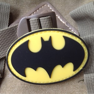 Könnte beinhalten: Ein ovaler Aufnäher mit dem schwarzen Batman-Logo, das auf einem gelben Hintergrund mit schwarzem Rand zentriert ist. Der Aufnäher ist entlang der Kante mit gelbem Faden vernäht und an einem braunen Stoffriemen mit einer Kunststoff-Verstellschnalle befestigt.