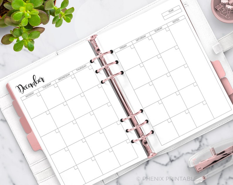 May include: A white monthly planner with a black and white grid for December. The planner has a pink tab for the month and a black and silver binder.