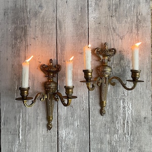Pode incluir: Duas arandelas de parede antigas de latão com velas brancas acesas. Cada arandela tem um design decorativo e comporta duas velas. As arandelas são montadas em um fundo de madeira cinza envelhecida, criando uma estética vintage.