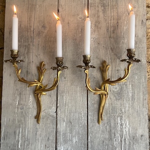 Pode incluir: Um par de arandelas de parede em metal dourado, cada uma com dois braços curvos com detalhes ornamentais em forma de folha. Cada arandela sustenta duas velas brancas, com todas as quatro velas acesas, emitindo um brilho quente.