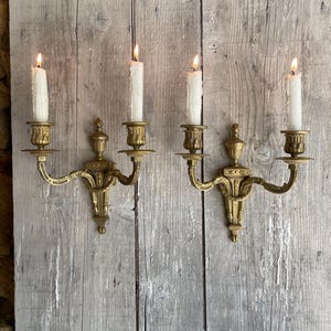 Pode incluir: Duas arandelas de latão antigas com velas brancas acesas. As arandelas estão montadas numa parede de madeira envelhecida. Cada arandela tem duas velas e apresenta detalhes ornamentados. As chamas são brilhantes e o latão tem uma pátina quente e envelhecida.