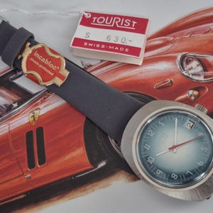 Peut inclure: Une montre-bracelet vintage avec un boîtier argenté et un cadran bleu, avec une trotteuse rouge et une fenêtre de date. La montre a un bracelet gris foncé et une étiquette "TOURIST" et "SWISS-MADE". Le fond comprend une voiture rouge.