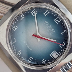 Peut inclure: Une montre-bracelet vintage avec un cadran dégradé bleu et argenté. La montre présente des chiffres blancs, des aiguilles blanches et une trotteuse rouge. Le cadran indique "TOURIST 17 JEWELS" et "INCABLOC". Le boîtier est argenté et le bracelet beige clair.