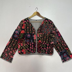 May include: A patterned jacket with a black base and a floral design in pink, red, and blue. The jacket features long sleeves and a V-neck with a striped lining. It is displayed on a wooden hanger.
