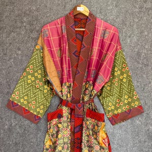 May include: A colorful, patterned robe with a belt. The robe features a mix of pink, green, brown, and red fabrics. The sleeves have a green geometric design, while the body has a pink plaid pattern. The robe is hanging on a wooden hanger.