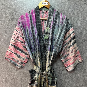May include: A long-sleeved kimono robe featuring a tie-dye pattern in shades of pink, purple, grey, and black. The garment includes a matching fabric belt at the waist and a dark grey collar. A small white tag labeled K.G.N is attached to the hanger.