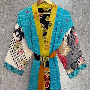 Może przedstawiać: Kolorowa szata w stylu kimono z patchworkowym wzorem. Szata ma turkusowy korpus z żółtymi wykończeniami i brązowym paskiem. Rękawy mają kwiatowe i geometryczne wzory. Szata wisi na drewnianym wieszaku.