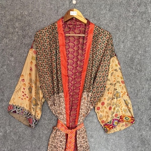 May include: A colorful, patterned kimono robe with a belt. The robe features a mix of floral, paisley, and geometric designs in shades of orange, red, beige, and green. The robe is hanging on a wooden hanger against a gray background.