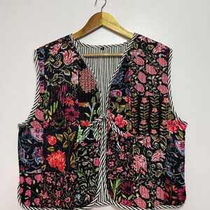 May include: A sleeveless quilted vest featuring a floral design in pink, red, and green on a black backdrop. The vest has a striped lining and trim, with a tie closure. It is displayed on a wooden hanger.