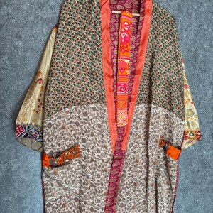 May include: A colorful kimono robe with a floral and paisley pattern. The robe features a mix of colors, including orange, pink, green, and cream. The robe is hanging on a wooden hanger against a gray background.