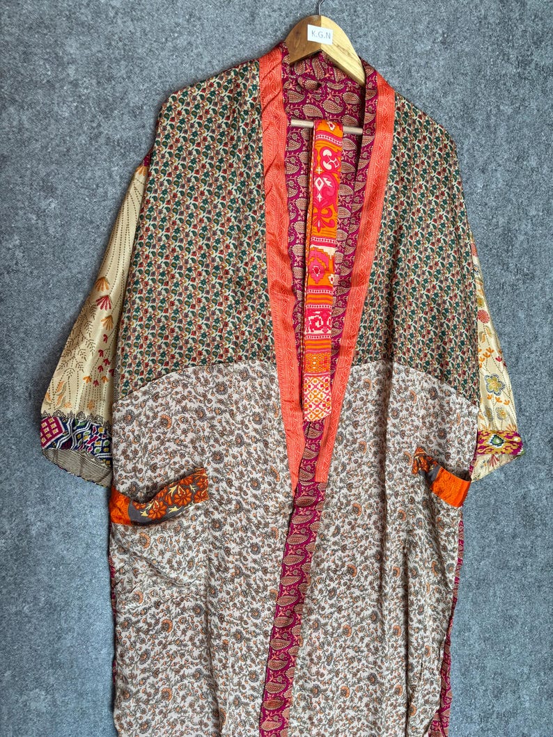 May include: A colorful kimono robe with a floral and paisley pattern. The robe features a mix of colors, including orange, pink, green, and cream. The robe is hanging on a wooden hanger against a gray background.