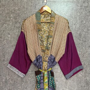 May include: A colorful, patterned kimono robe with long sleeves. The robe features a mix of geometric and floral patterns in shades of purple, tan, green, and blue. The sleeves are a solid purple with a patterned border. The robe is hanging on a wooden hanger.