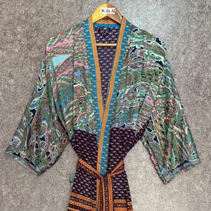 May include: A colorful paisley print kimono robe with a tie belt. The robe features a mix of blue, green, pink, and orange patterns. The robe is hanging on a wooden hanger with the letters "K.G.N" on it.