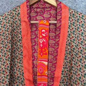 May include: A colorful kimono with a floral pattern in shades of green, red, and cream. The interior lining is a deep pink with a paisley design. The edges are trimmed with a bright orange border. A matching orange sash hangs in the center.