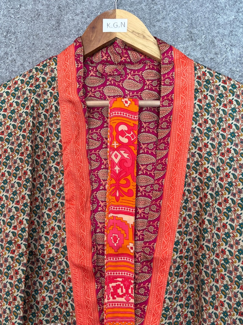 May include: A colorful kimono with a floral pattern in shades of green, red, and cream. The interior lining is a deep pink with a paisley design. The edges are trimmed with a bright orange border. A matching orange sash hangs in the center.