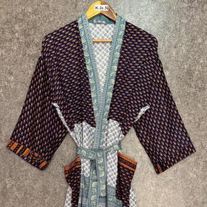 Pode incluir: Robe estilo quimono azul marinho com um padrão repetido de pequenas formas laranja. O robe tem uma borda estampada em azul claro e branco e um cinto a condizer. O robe está pendurado em um cabide de madeira.