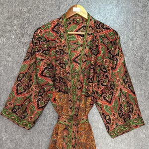 Pode incluir: Robe com padrão paisley com cinto, pendurado em um cabide de madeira. O robe apresenta uma mistura de cores vermelha, verde, preta e dourada. O robe tem mangas compridas e um design envolvente, perfeito para relaxar.