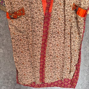 May include: A beige kimono-style robe with a floral pattern. The robe features orange trim along the edges and pockets, and a paisley border. The fabric appears soft and lightweight, suitable for layering or lounging.