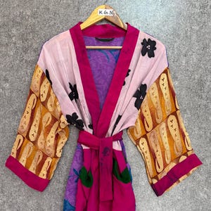 Op de afbeelding: Een kleurrijke, bedrukte kimono met een strikceintuur. De kimono heeft een mix van roze, paarse en oranje stoffen met bloemen- en paisley-ontwerpen. Zwarte bloemaccenten sieren de lichtroze stof. De kimono hangt aan een houten kleerhanger.