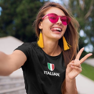 May include: A woman wearing a black t-shirt with a white Italian flag and the text "ITALIAN." She is wearing pink sunglasses and yellow tassel earrings. She is smiling and giving a peace sign.