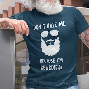 Puede incluir: Una camiseta azul marino con un gráfico blanco de un hombre barbudo con gafas de sol. El texto en la camiseta dice "Don't hate me because I'm Beardiful."