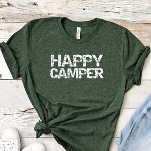 Puede incluir: Una camiseta verde con el texto "HAPPY CAMPER" en letras blancas desgastadas. La camiseta est&aacute; colocada sobre un fondo de madera blanca con un par de zapatillas blancas y unos vaqueros azules.