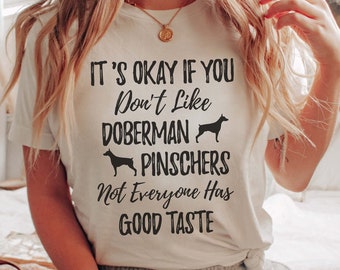 Op de afbeelding: Wit T-shirt met zwarte tekst die luidt: "It's okay if you don't like Doberman Pinschers. Not everyone has good taste." Twee zwarte silhouetten van Dobermann Pinschers zijn opgenomen in de tekst.