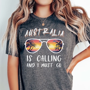 Może przedstawiać: Szary T-shirt z nadrukiem "Australia is calling and I must go". Tekst jest otoczony parą okularów przeciwsłonecznych z odbitym w szkle zachodem słońca i palmami.