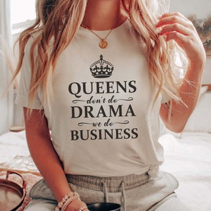 May include: A cream-coloured short-sleeve t-shirt featuring a black graphic of a crown above the text "QUEENS don't do DRAMA we do BUSINESS" in a mix of serif and script fonts. The shirt is paired with grey sweatpants and a black mug.