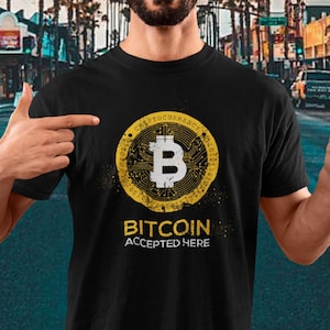 May include: A man wearing a black t-shirt with a gold Bitcoin logo and the text "Bitcoin Accepted Here". He is smiling and holding a smartphone with a Bitcoin logo on the screen.