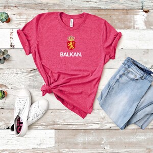 K&ouml;nnte beinhalten: Ein pinkfarbenes T-Shirt mit einer wei&szlig;en Grafik eines roten Schildes mit einem L&ouml;wen und einer Krone sowie dem Wort "BALKAN." Das Shirt liegt auf einer wei&szlig;en Holzoberfl&auml;che mit einem Paar wei&szlig;en Sneakers und einer blauen Jeans.