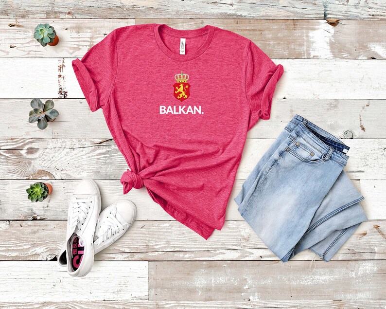 K&ouml;nnte beinhalten: Ein pinkfarbenes T-Shirt mit einer wei&szlig;en Grafik eines roten Schildes mit einem L&ouml;wen und einer Krone sowie dem Wort "BALKAN." Das Shirt liegt auf einer wei&szlig;en Holzoberfl&auml;che mit einem Paar wei&szlig;en Sneakers und einer blauen Jeans.