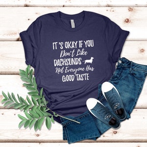 May include: A navy blue t-shirt with white text that reads "It's okay if you don't like dachshunds. Not everyone has good taste." A white dachshund silhouette is next to the text.