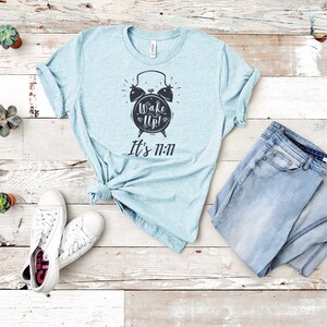 May include: A light blue t-shirt with a black alarm clock graphic and the text "Wake Up! It's 11:11". The shirt is laid out on a white wooden surface with a pair of white sneakers and blue jeans.