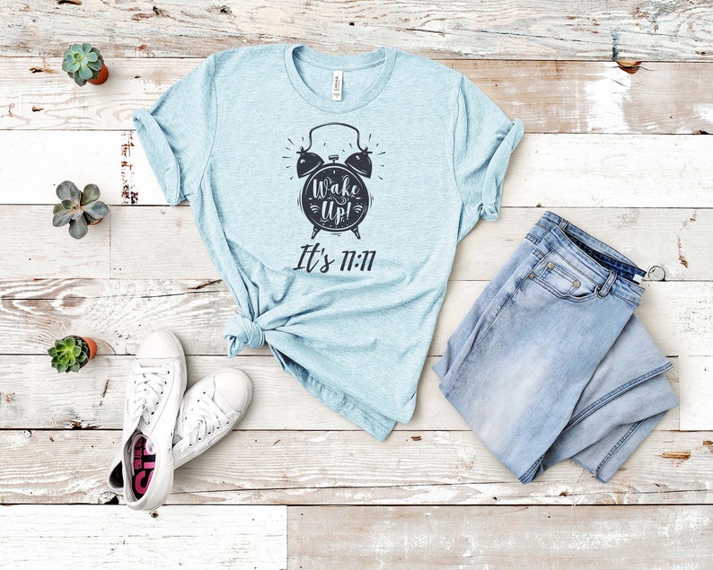 May include: A light blue t-shirt with a black alarm clock graphic and the text "Wake Up! It's 11:11". The shirt is laid out on a white wooden surface with a pair of white sneakers and blue jeans.
