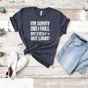 May include: A navy blue t-shirt with the text "I'M SORRY DID I ROLL MY EYES OUT LOUD?" in white. The shirt is paired with light blue jeans and white sneakers. The shirt is knotted at the waist.