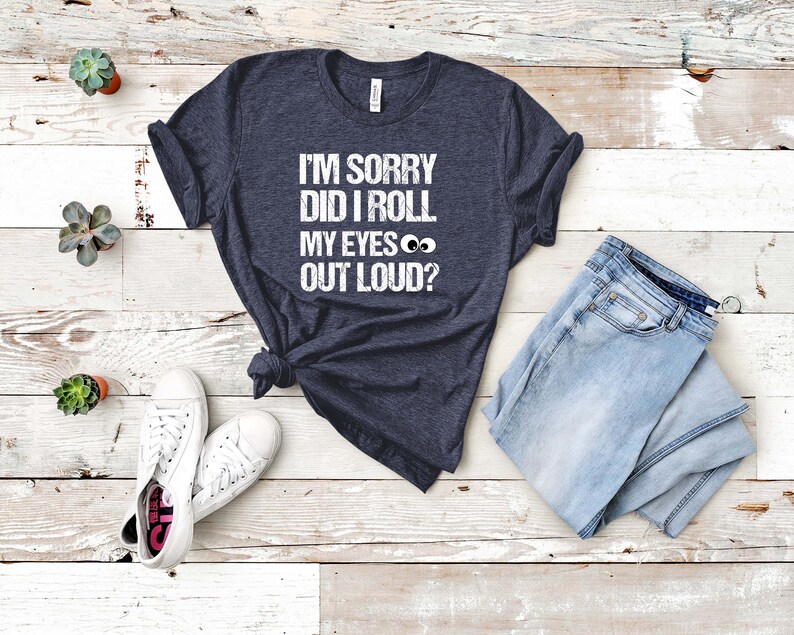 May include: A navy blue t-shirt with the text "I'M SORRY DID I ROLL MY EYES OUT LOUD?" in white. The shirt is paired with light blue jeans and white sneakers. The shirt is knotted at the waist.