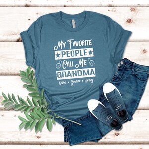 May include: A teal blue t-shirt with the text "My Favorite People Call Me Grandma" in white with stars and thumbs up. The names "Evan, Gunner, Jenny" are printed below the text.