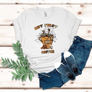 May include: White t-shirt featuring a skeleton sitting in a coffee cup with a spider web and the text "But First Coffee".