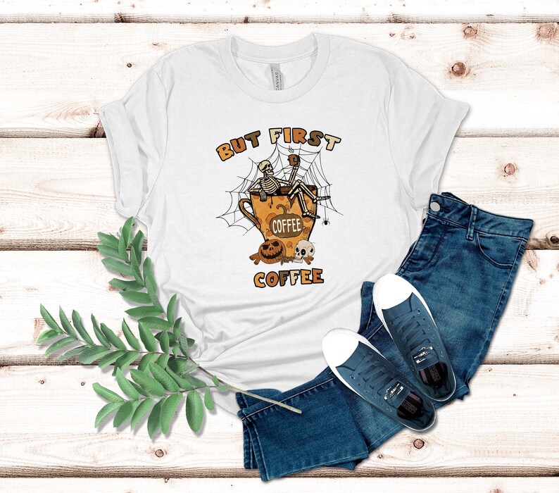 May include: White t-shirt featuring a skeleton sitting in a coffee cup with a spider web and the text "But First Coffee".