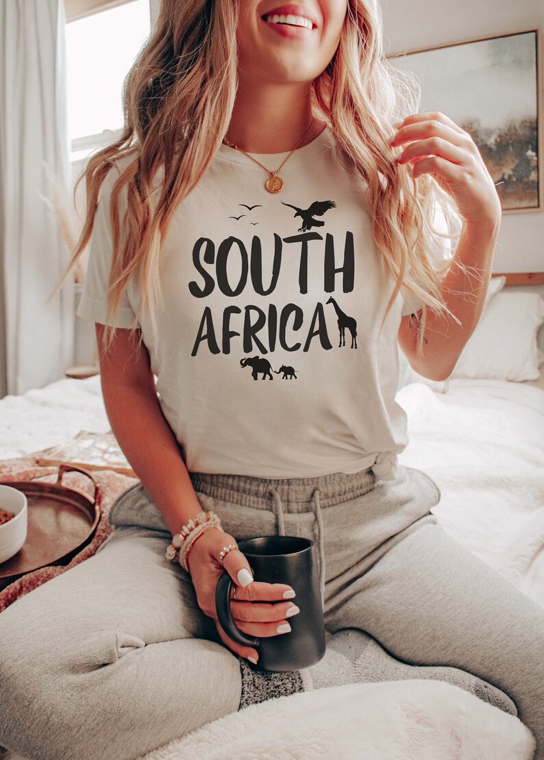 May include: Cream-colored t-shirt with the words "SOUTH AFRICA" in bold black letters. The design includes silhouettes of birds, an eagle, a giraffe, and elephants. The person is wearing gray sweatpants and holding a black mug.