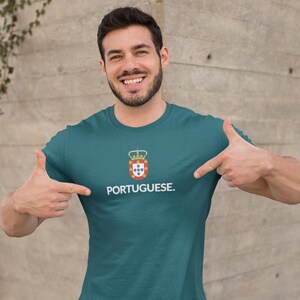 Może przedstawiać: Turkusowa koszulka z białym napisem "PORTUGUESE". Nad słowem znajduje się herb z koroną i tarczą. Osoba na zdjęciu wskazuje na koszulkę.