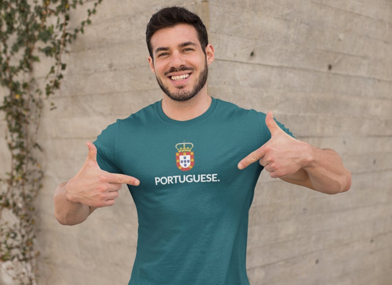 Może przedstawiać: Turkusowa koszulka z białym napisem "PORTUGUESE". Nad słowem znajduje się herb z koroną i tarczą. Osoba na zdjęciu wskazuje na koszulkę.