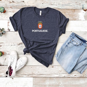 Może przedstawiać: Granatowa koszulka z napisem "PORTUGUESE" w kolorze białym, pod herbem z czerwonymi, ż&oacute;łtymi i niebieskimi akcentami. Koszulka jest zestawiona z jasnoniebieskimi dżinsami i białymi trampkami.